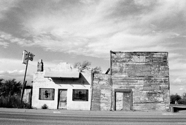 A black and white photo of two dilapidated buildings, one brick and one wood, with boarded up windows and a crooked sign, stand on a roadside.