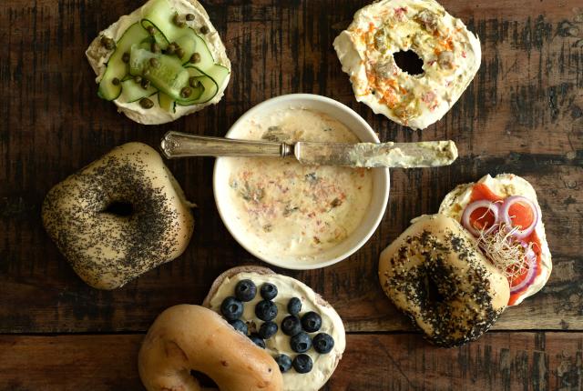 An assortment of bagels with various toppings and a bowl of cream cheese sit on a wooden surface.