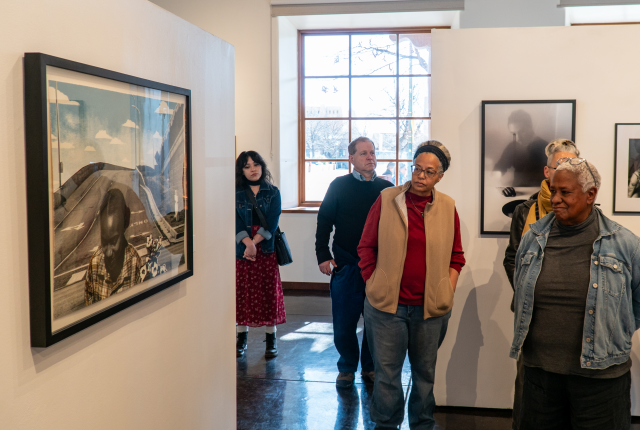 A diverse group of people attentively observe framed photographs in a well-lit art gallery.