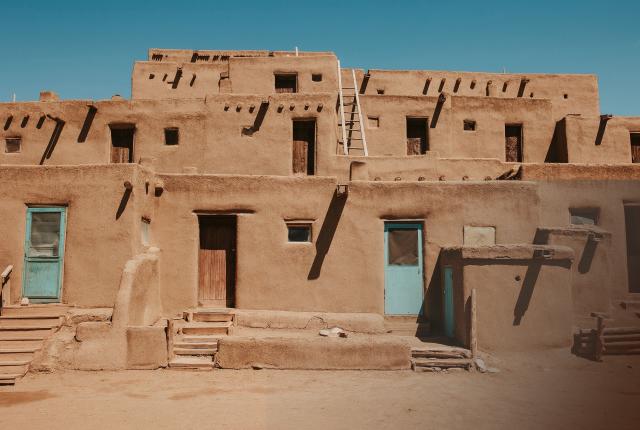 A multi‑story adobe dwelling with tan earthen walls, wooden doors, small square windows, and ladders stands under a clear blue sky.