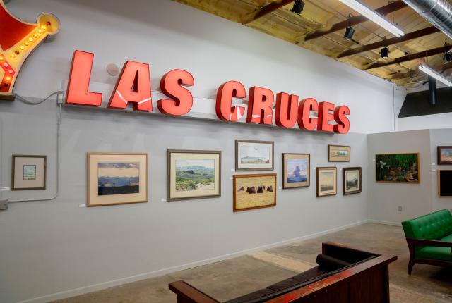 A bright red vintage sign reading "Las Cruces" is mounted above a collection of framed landscape paintings on a gallery wall.