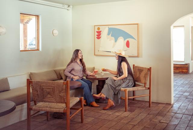Two women sit at a round table in a cozy, minimalist room with ceramic floor tiles.