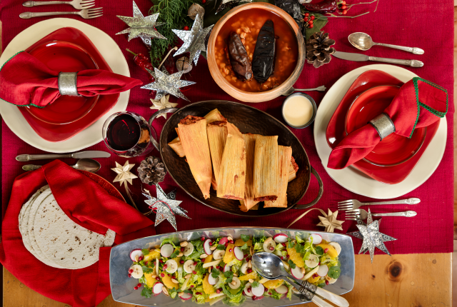 An overhead view of a holiday table set with red place settings, tamales, salad, and pozole.