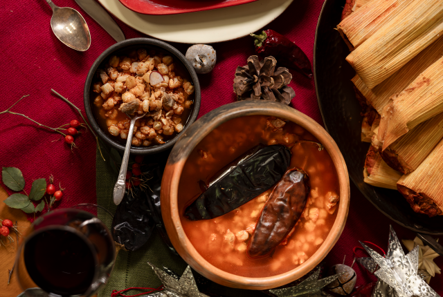 A festive table with traditional Mexican dishes, including pozole, tamales, and a glass of red wine, with holiday decorations scattered around.