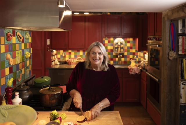 Cheryl Alters Jamison smiles while cutting fruit on a wooden cutting board in a colorful kitchen.