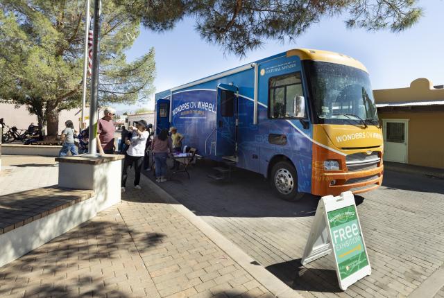 Mobile museum bus parked on a plaza with visitors gathered near a sign advertising a free exhibition.