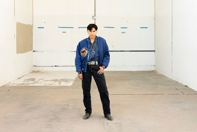Jojo Jackson in a blue jacket with a beaded design, black jeans, and boots stands in a concrete room.