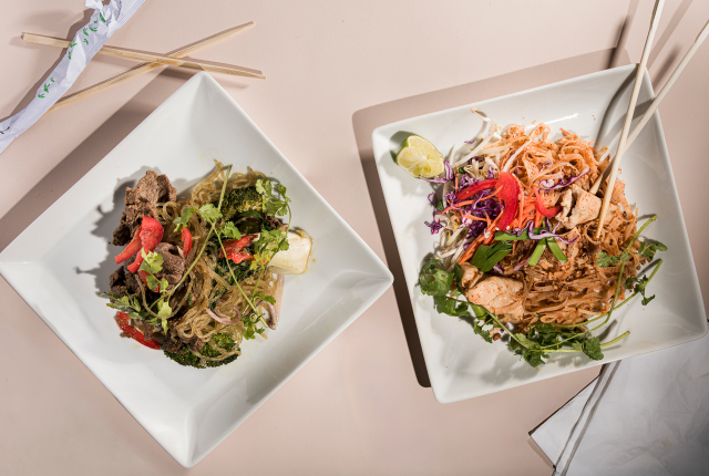 Two square plates on a light surface; left dish has beef and vegetables, right has pad thai with chicken.