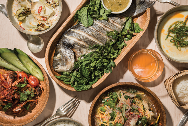 An array of Thai dishes on a table: whole fish with herbs, spicy salad, soup, and grilled vegetables.