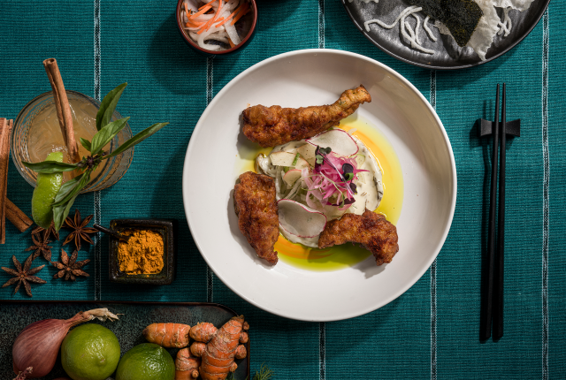 A white plate with crispy fried chicken topped with radish slices and microgreens, set against a teal cloth. Ingredients and chopsticks surround it.