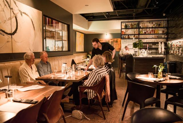 A cozy, modern restaurant with dim lighting. A server tends to four older individuals seated at a wooden table.