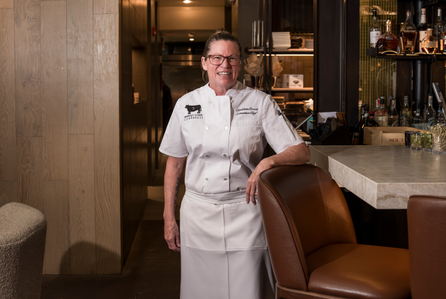 Chef Kathleen Crook in a white uniform stands smiling beside a marble bar with brown leather chairs.