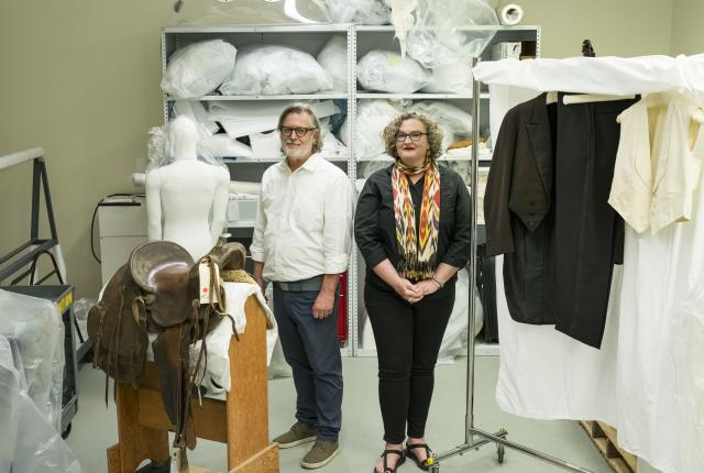 Bradford Epley and Angela Duckwall stand between a saddle and a rack of garments inside a museum conservation lab.