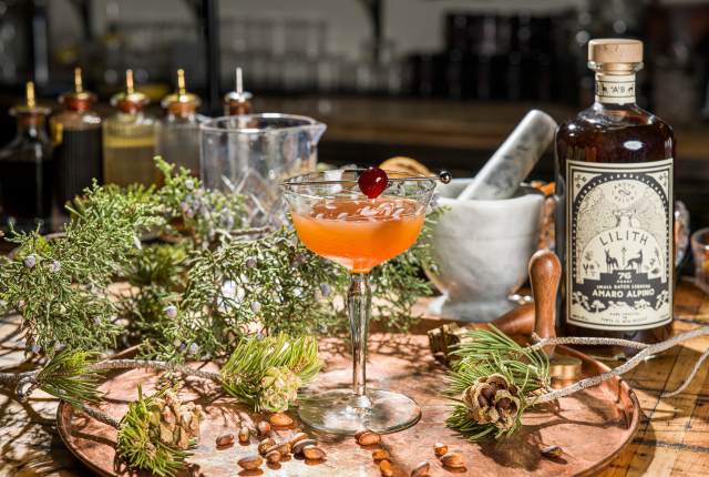 Amber cocktail in a stemmed glass surrounded by herbs, spices, and a mortar and pestle.