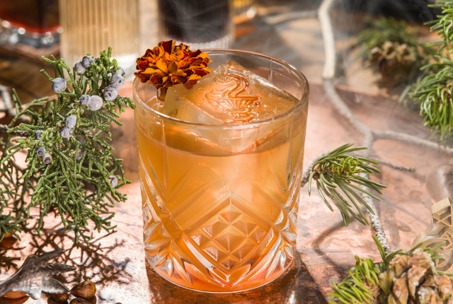 Orange cocktail with a flower garnish and a large patterned ice cube, surrounded by juniper and pine.