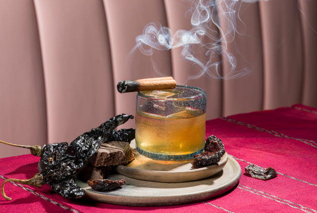 Amber cocktail with smoking garnish, surrounded by dried red chiles on a red tablecloth.