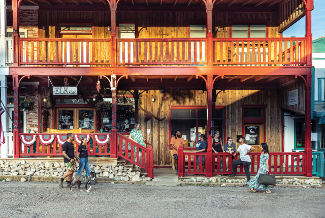 Two-story wooden building with red railings. People gather outside under warm sunlight; a couple walks two dogs, and others chat casually.