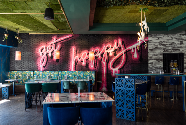 A brightly lit bar with a pink and white neon "Get Happy" sign, floral booths, and a faux-grass ceiling.