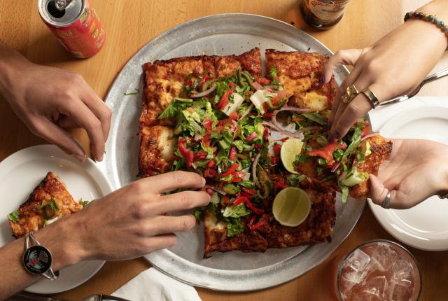 Hands grabbing slices of a rectangular pizza topped with fresh vegetables and lime wedges on a metal tray.