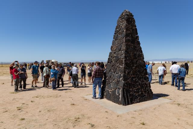 Group of visitors gathered around a tall stone monument in a desert landscape under a clear sky.