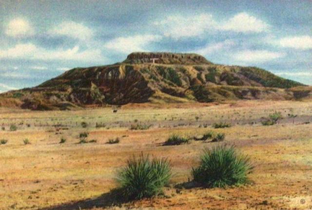 A flat-topped, rocky mesa rises above a dry desert plain dotted with low shrubs under a partly cloudy sky.