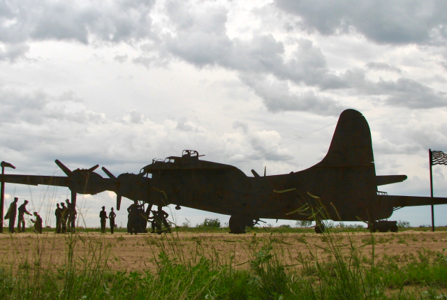 A silhouette of a bomber plane sits on a grassy field with a line of people figures standing nearby.