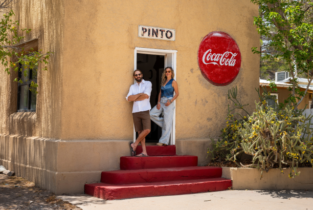 Two individuals stand in the doorway of a tan stucco building with a “PINTO” sign above the door and a large vintage Coca‑Cola sign on the wall, with red steps and cactus plants in front.