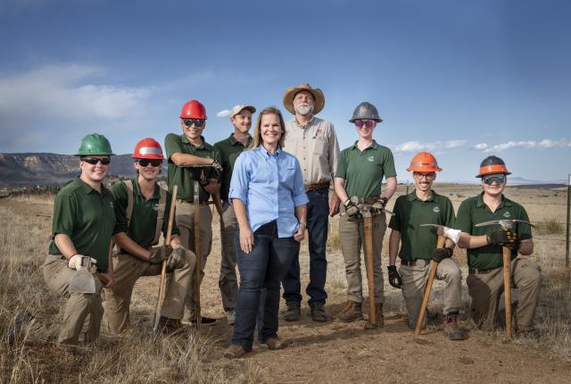 A group of people in outdoor work gear and hard hats pose together in a grassy open landscape, holding tools and equipment.