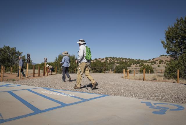 People with hats and backpacks walk along a sunny trailhead in a desert landscape with scattered trees and hills.