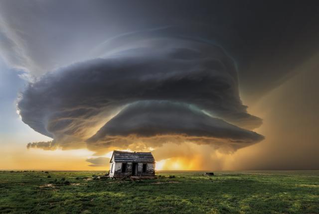 A towering supercell storm glows at sunset over eastern New Mexico grasslands near an abandoned farmhouse.
