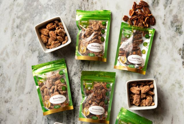 Assorted bags of flavored pecans from Worthington Farms, including butter pecan, red chile, and blueberry, arranged on a marble surface beside small bowls of candied pecans.