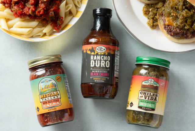 Three jars of condiment are displayed on a table: salsa, barbecue sauce, and green chiles. Nearby are pasta and a burger on plates.