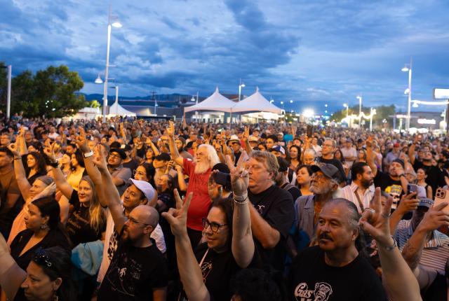 A large crowd at an outdoor event raises hands and phones toward a stage at dusk, with tents, lights, and a cloudy blue sky in the background.