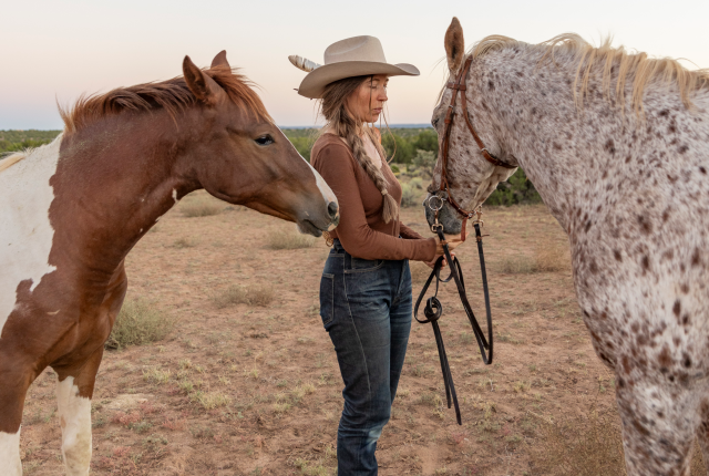 Artist Stella Maria Baer stands between two horses in the Santa Fe countryside, wearing a hat and holding a rope.