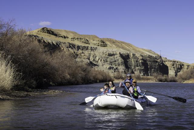 A family enjoys rafting on a calm river surrounded by barren hills and dry brush under a clear blue sky.