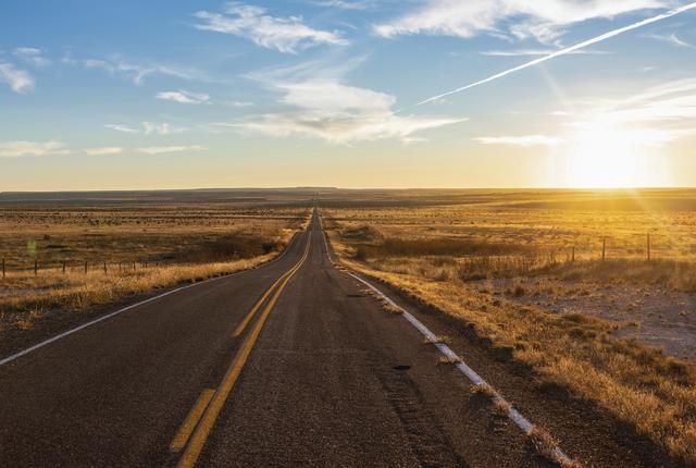 A two-lane road stretches toward the horizon through golden grassland beneath a blue and yellow sky.