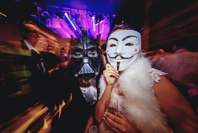 Two partygoers wearing Darth Vader and Guy Fawkes masks beneath purple lights.