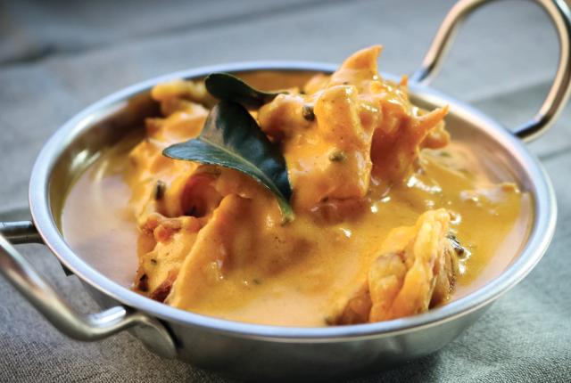 A bowl of kadhi pakora curry featuring creamy yellow yogurt-based sauce with visible spices and pieces of chickpea flour dumplings.