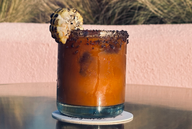Orange cocktail with a dark salt rim, garnished with a charred corn slice.