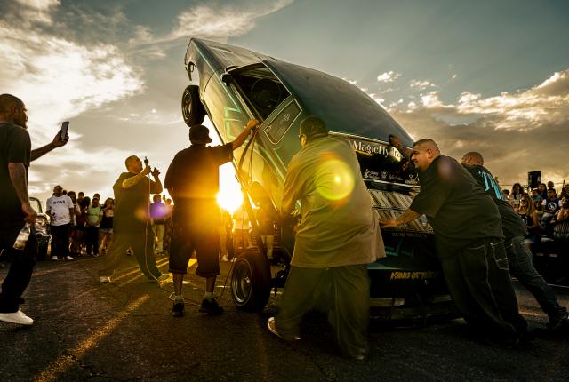 Two Taking Over Car Club members brace the rear of a lowrider as another man operates the switches, causing the car to hop during a crowded Duke Day event.