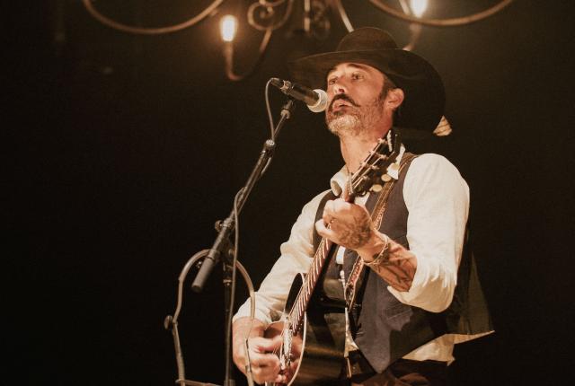 Ryan Bingham sings into a microphone while playing an acoustic guitar.