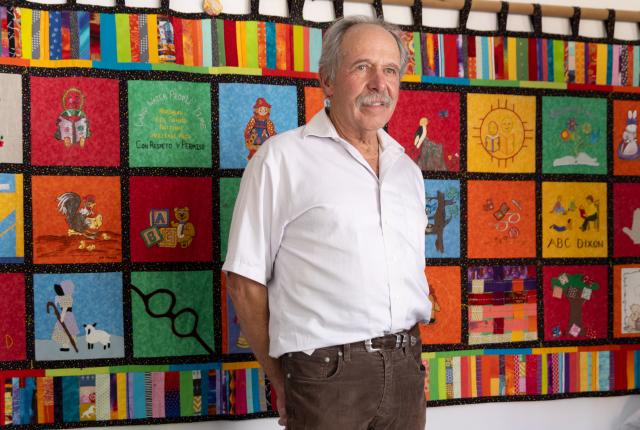 Man in white shirt and brown pants stands smiling in a room. Behind him, a colorful quilt with various panels hangs on the wall.
