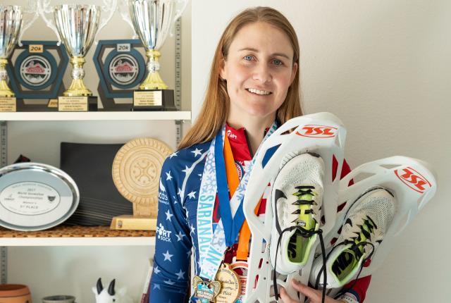 Michelle Hummel stands in front of her trophy shelf holding racing snowshoes and wearing medals.
