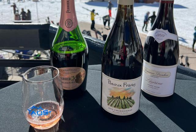 Three wine bottles and a glass on a table overlook a snowy ski resort during daylight.
