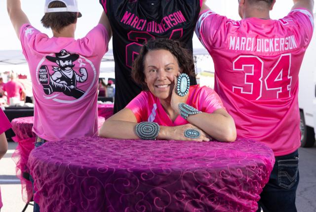 Woman smiles, leaning on a magenta table, wearing pink.
