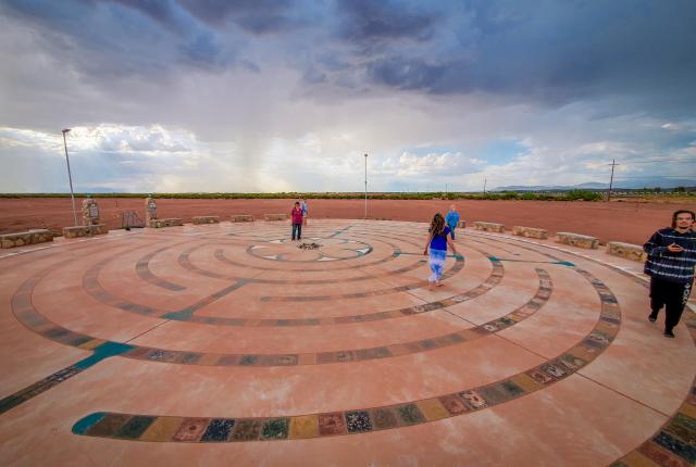 People walk on a circular labyrinth on a concrete surface under a dramatic, cloudy sky.