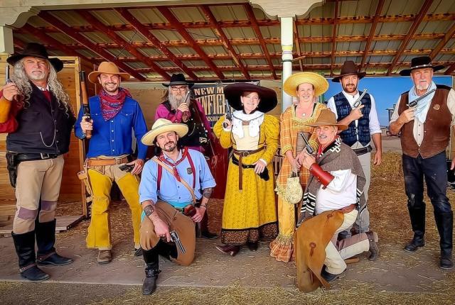 Group of people dressed as cowboys and cowgirls with guns posing under a wooden-roofed shelter in a western-themed setting.