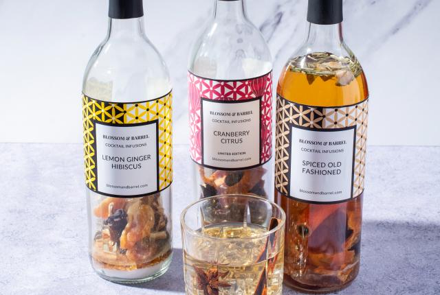 Three bottles of cocktail infusions labeled Lemon Ginger Hibiscus, Cranberry Citrus, and Spiced Old Fashioned stand on a marble surface, with a glass of infused drink in front.