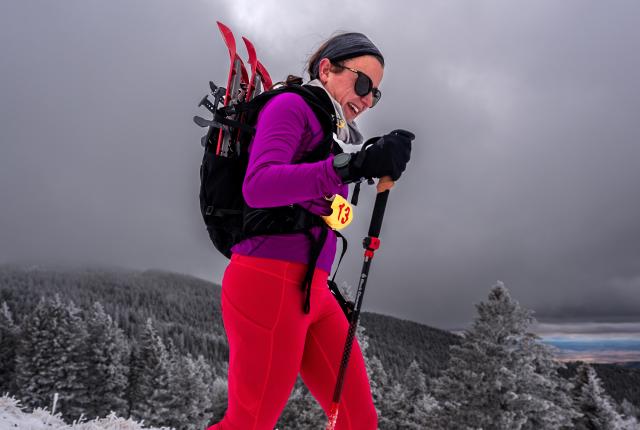A woman in vibrant red and purple gear hikes a snowy mountain with skis strapped to her backpack.