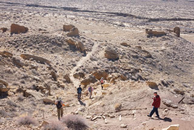 A group of people hike along a rugged trail in a vast desert landscape, surrounded by scattered rock formations.